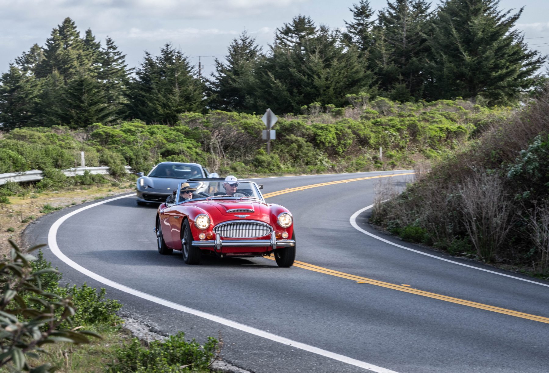 Members touring the coast road in an Austin-Healey, a Ferrari following behind